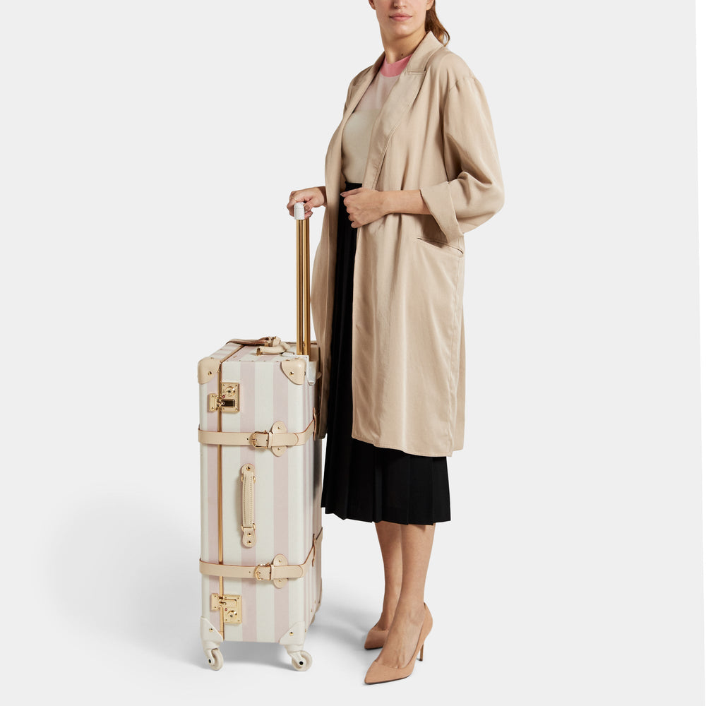 Three-quarter view of The Illustrator check-in spinner suitcase in pink check pattern with gold handle and spinner wheels
