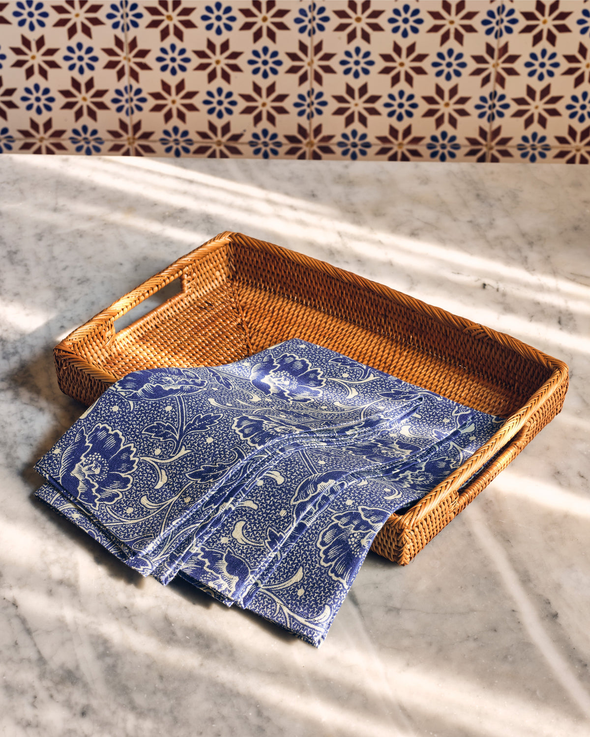 Folded front view of Toscana Blue linen napkins, set of 4, showing blue-and-white floral vine pattern draped in a woven rattan tray on a marble tabletop