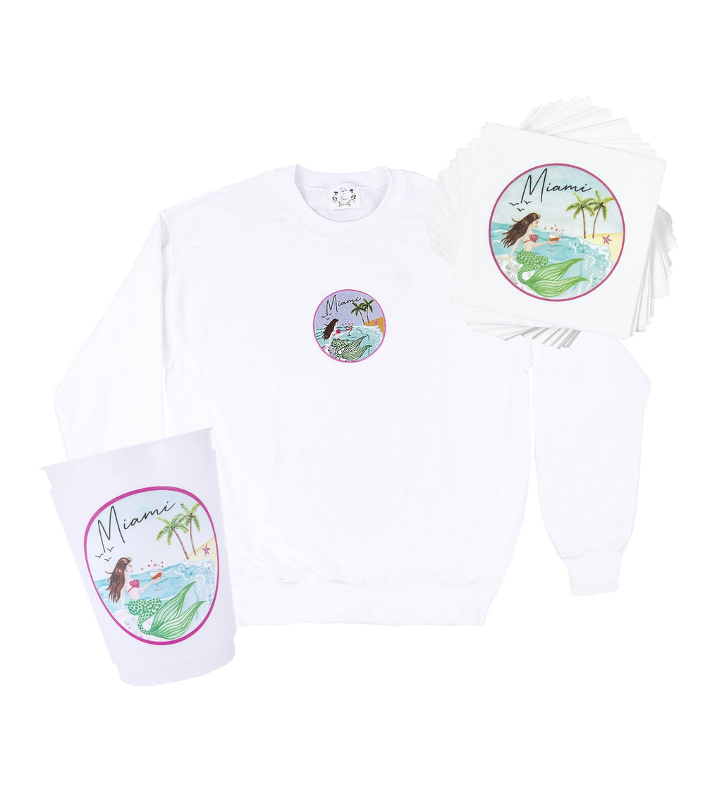 Front view of white crewneck sweatshirt, stack of napkins, and cup showcasing round Miami mermaid crest design on white background