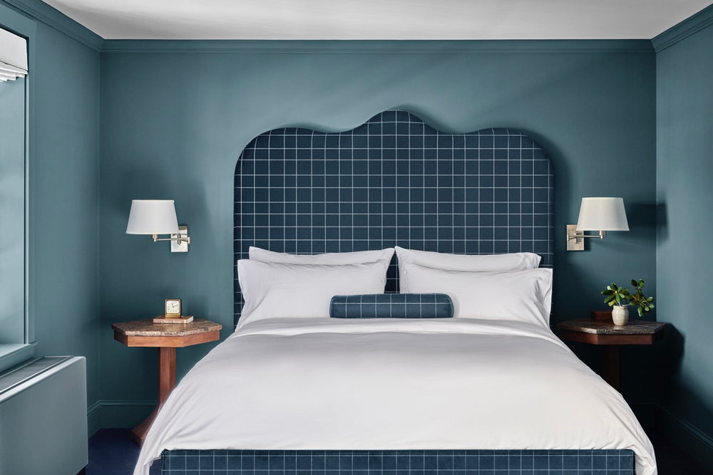 King bed centered in teal hotel room with white linens, checked navy headboard and bolster, wooden nightstands and wall sconces