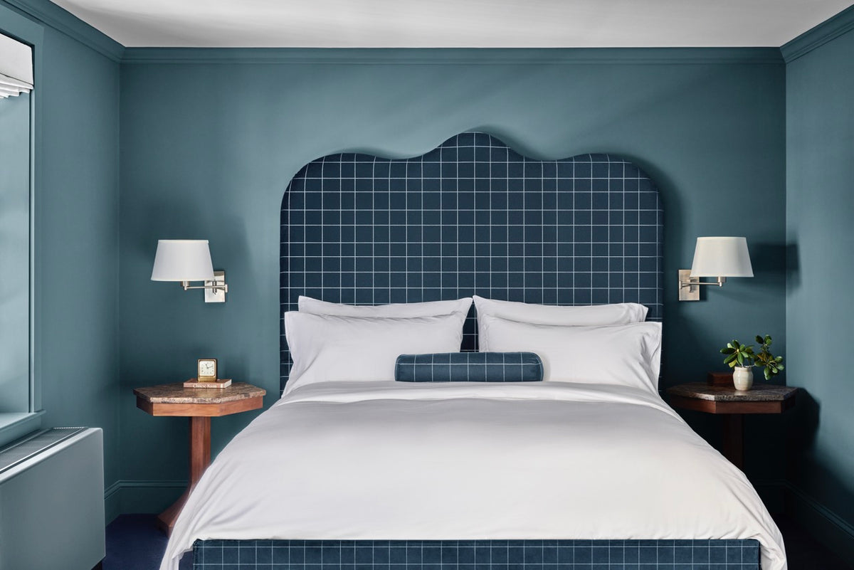King bed centered in teal hotel room with white linens, checked navy headboard and bolster, wooden nightstands and wall sconces