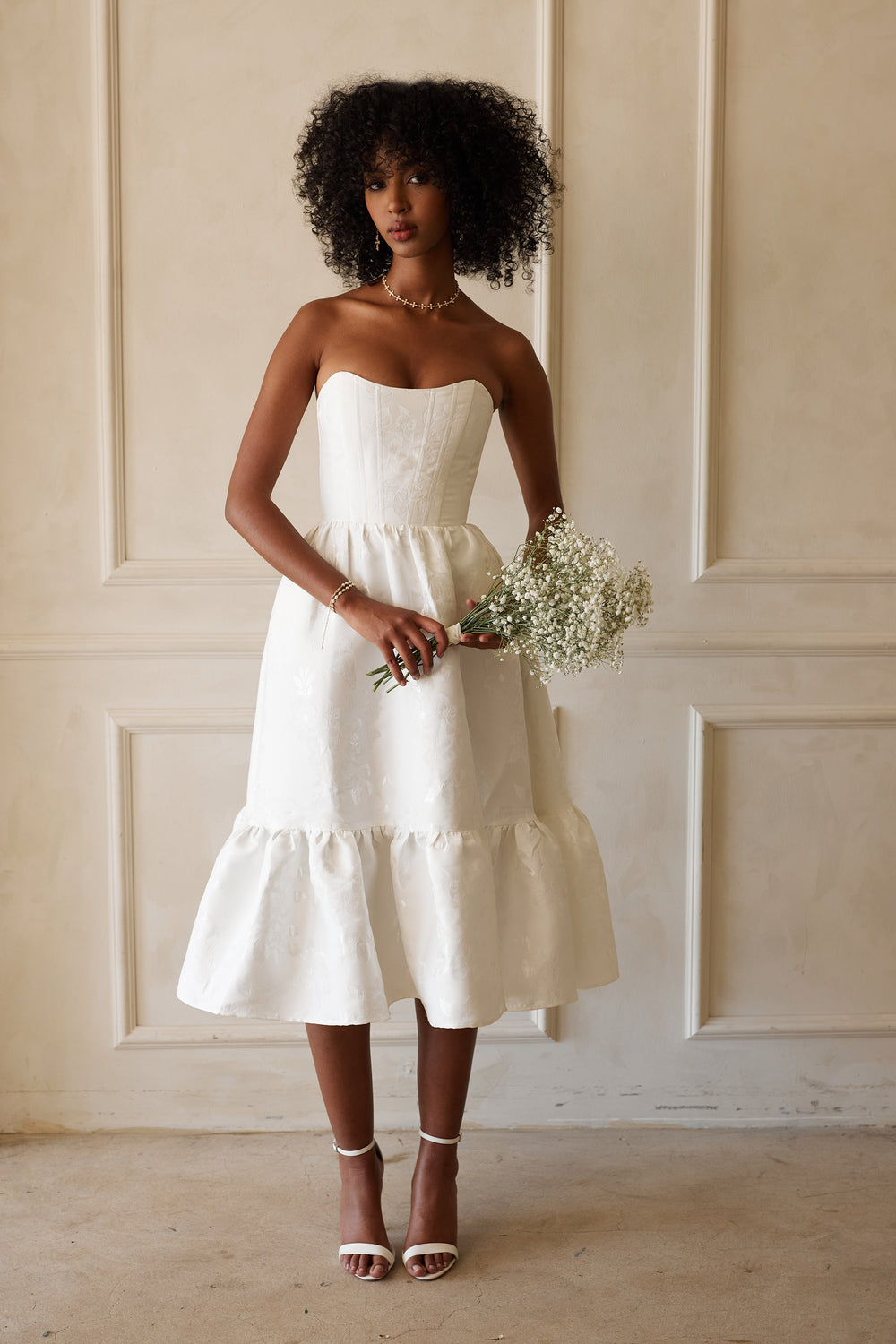 Front view of Virginia dress in white brocade floral with strapless corset bodice and ruffle hem against cream wall