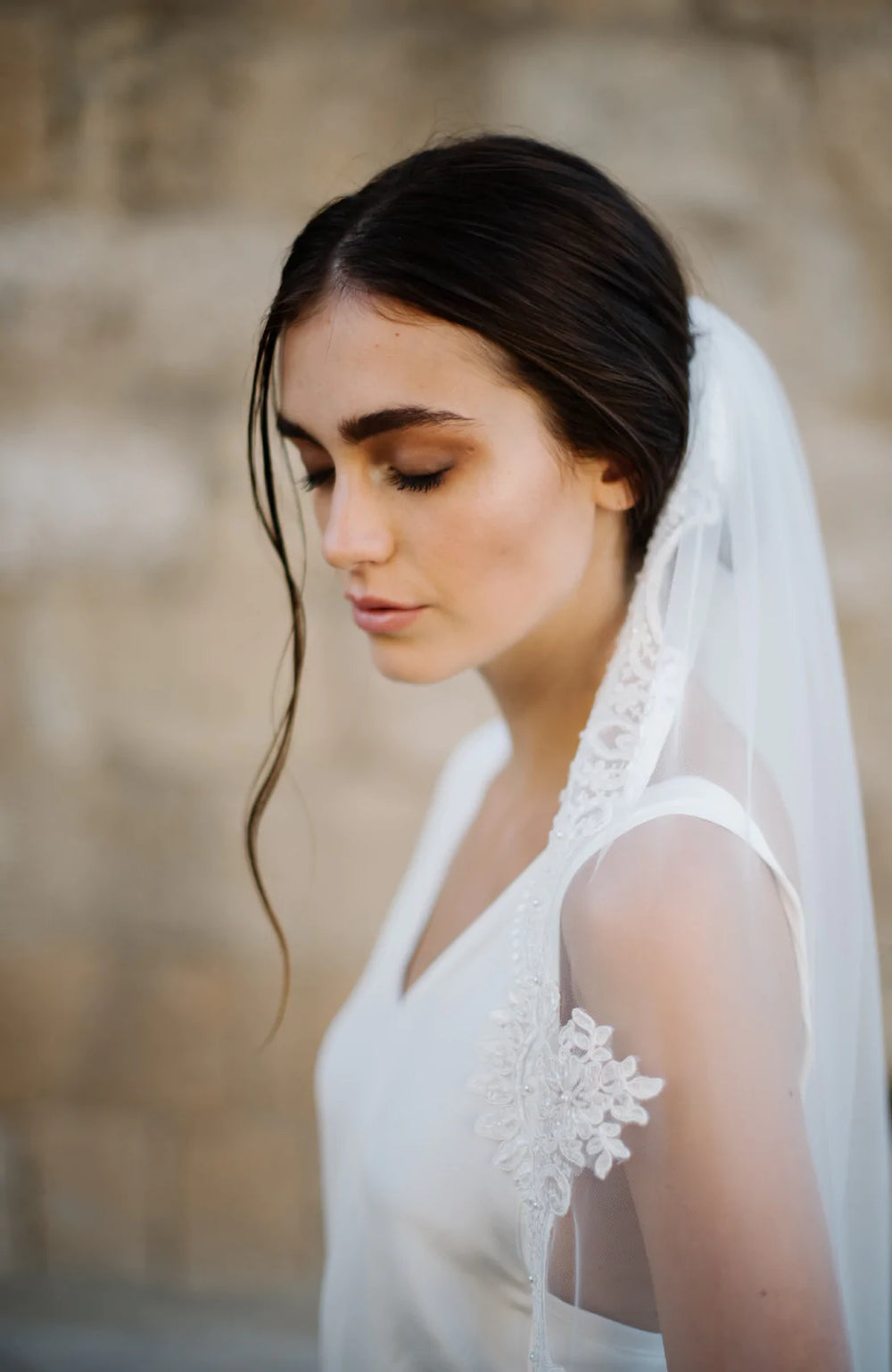 Side profile of model wearing Venetia lace veil showing lace applique and pearl beadwork against blurred stone background