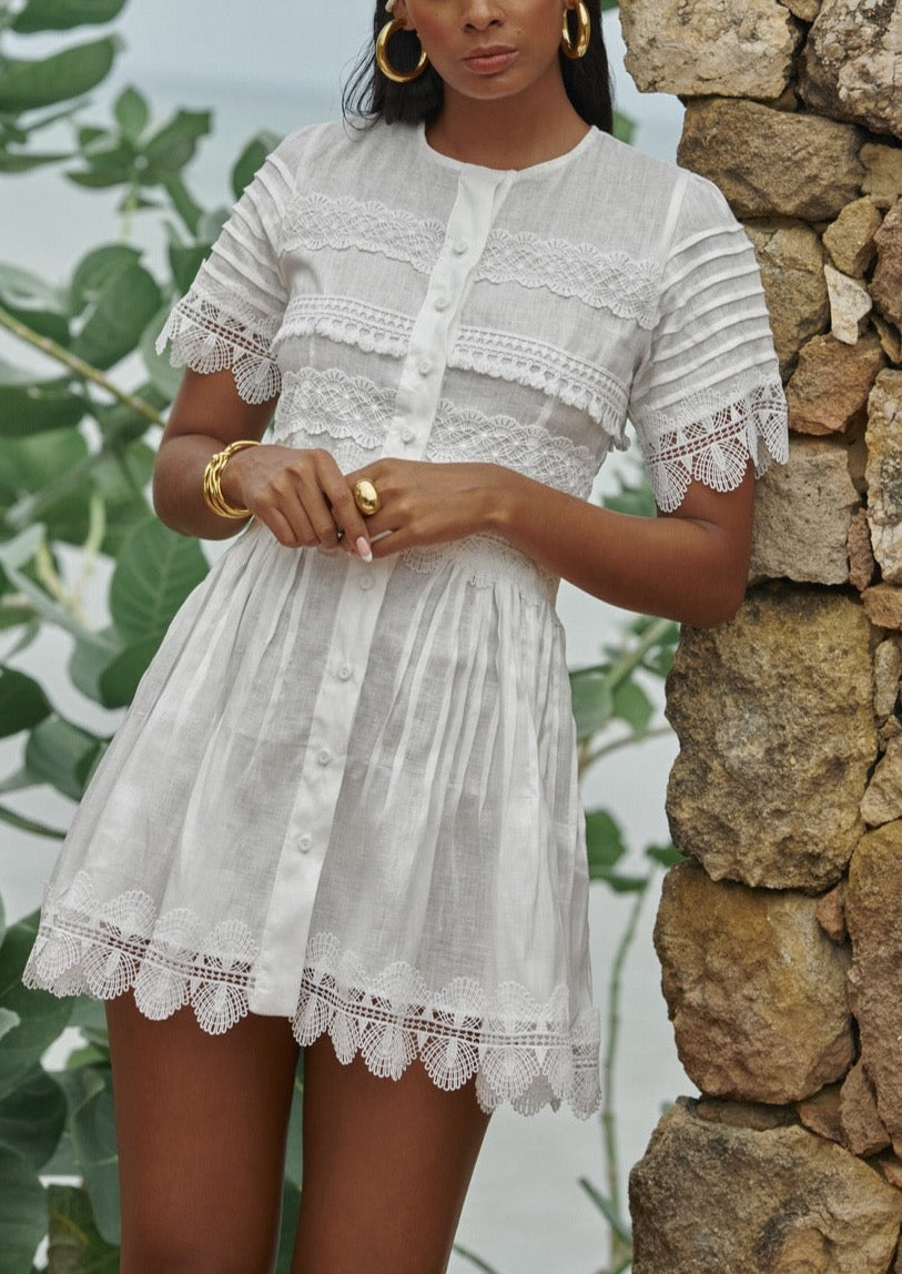 Front of Waimari Violetta white mini dress showing lace inserts, button-front closure and scalloped hem against stone wall