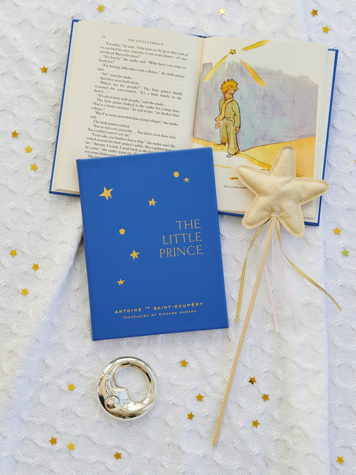 Flat lay of blue bonded leather The Little Prince book with gold-foil title, open illustration, and star wand on white blanket