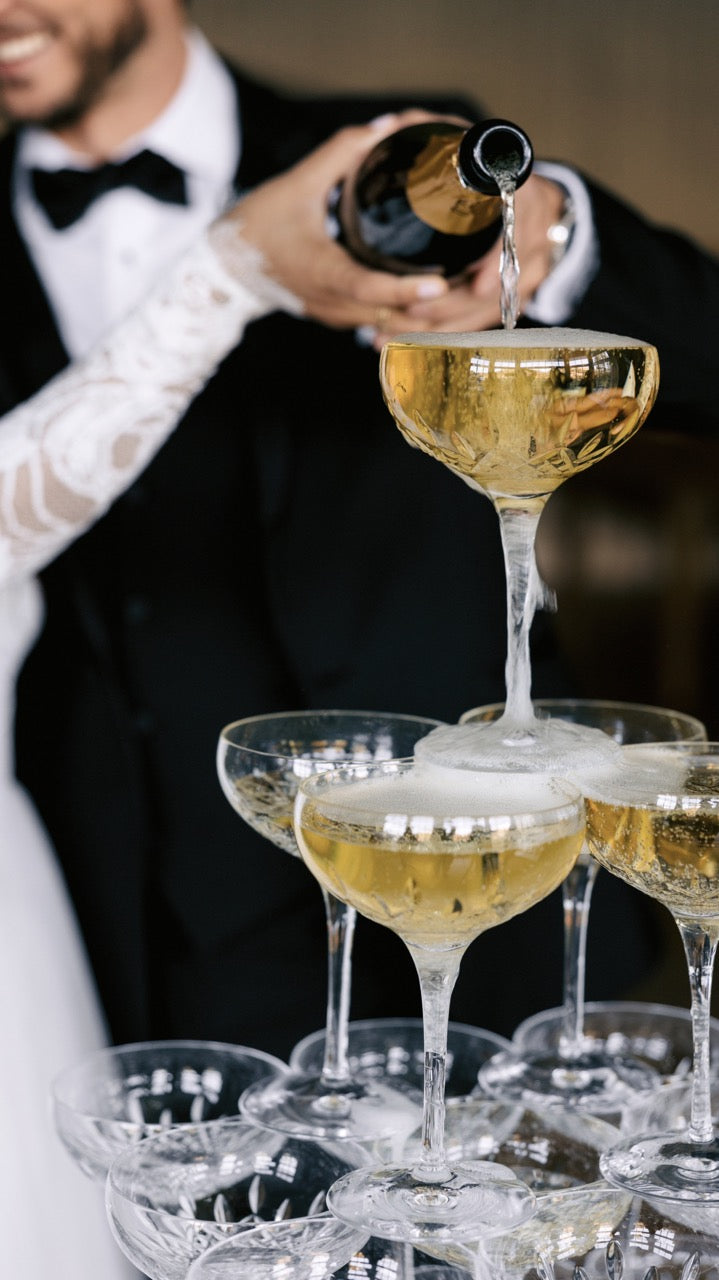 Bride and groom pouring champagne into a stacked tower of Lismore Essence 9.5 oz champagne saucers