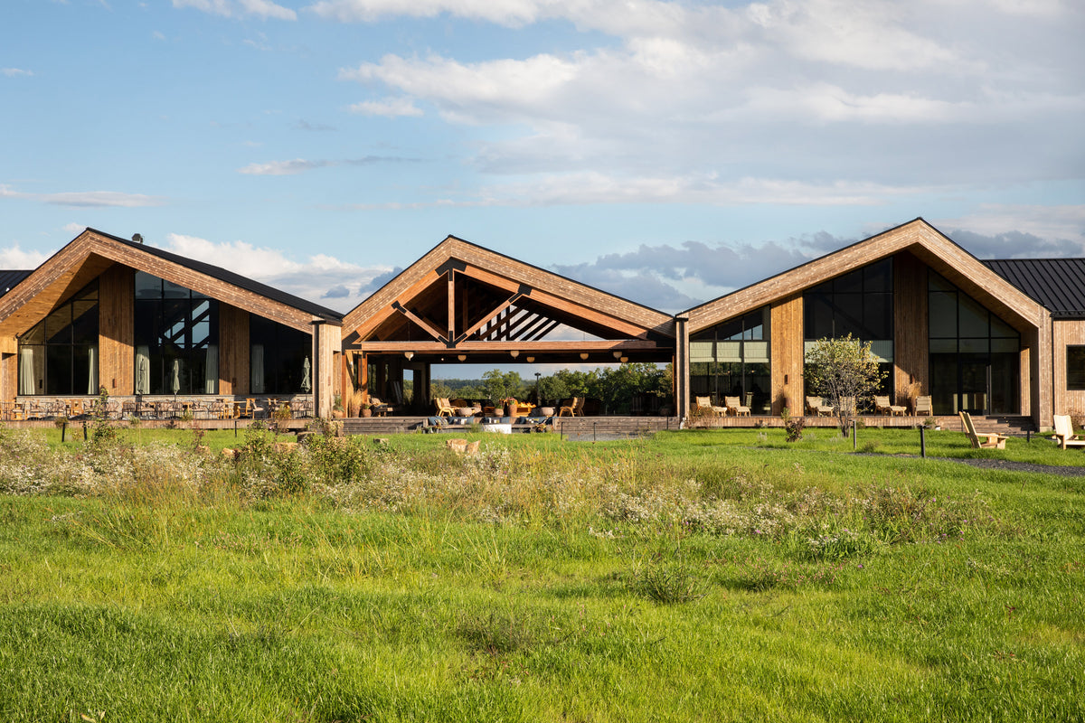 Exterior of Wildflower Farms lodge with three peaked wooden buildings, outdoor seating, and a grassy meadow under a blue sky