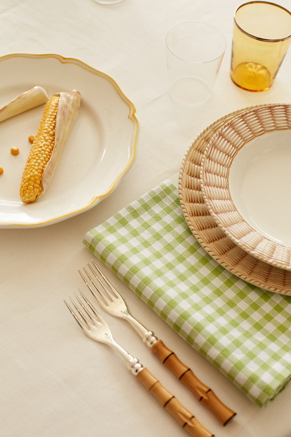 Top down view of Vichy napkin in sage gingham on a cream tablecloth beside a wicker rim plate and bamboo handled forks