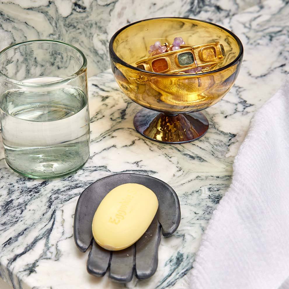 Iridescent amber glass footed bowl holding jewelry on a marble countertop beside a glass of water and a hand-shaped soap dish
