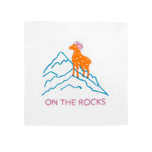 Top view of linen cocktail napkin with embroidered orange ram on blue mountains, the words ON THE ROCKS, and hemstitch border