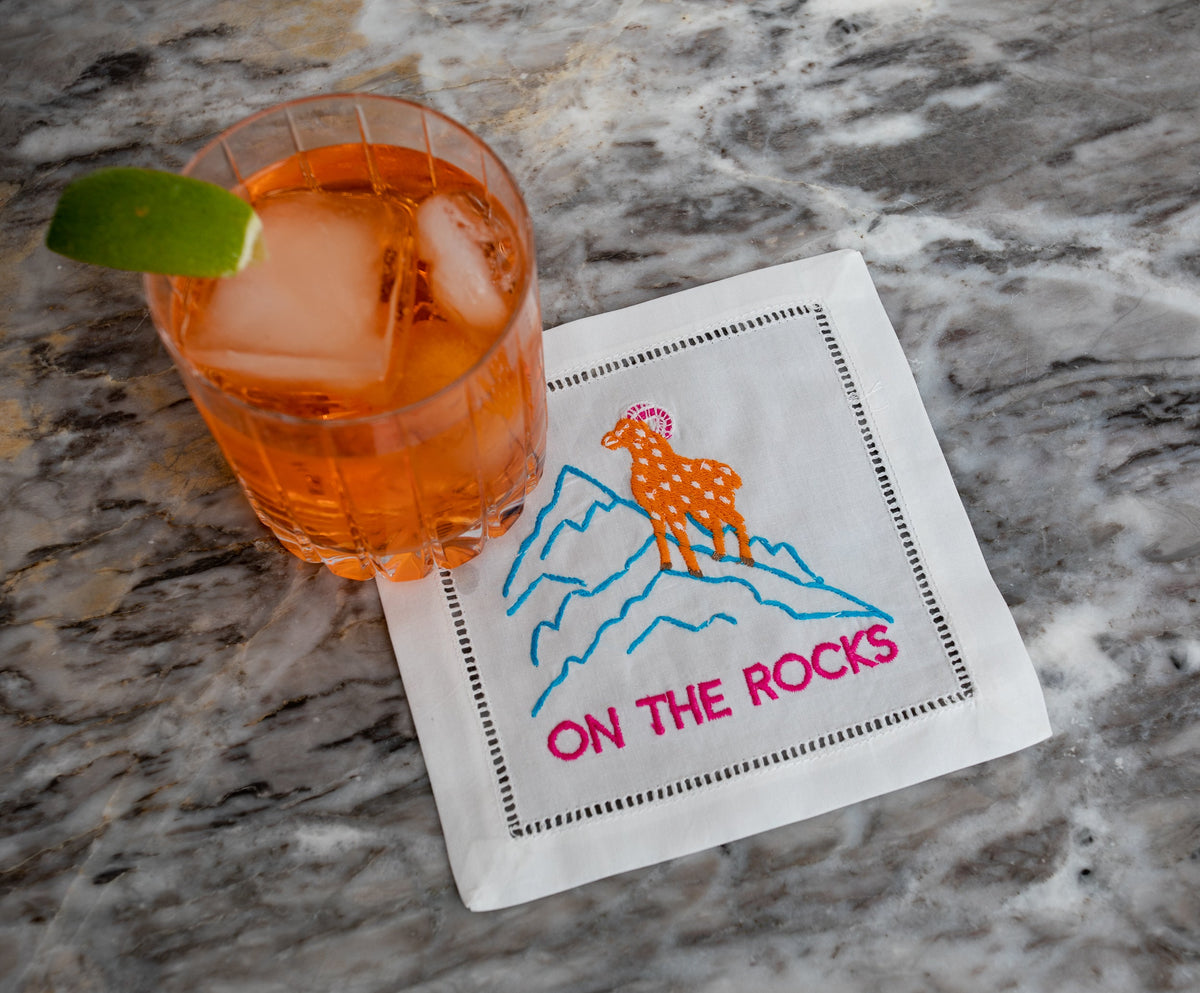 Angled view of On The Rocks cocktail napkin set showcasing an embroidered ram and mountains beside a cocktail on marble