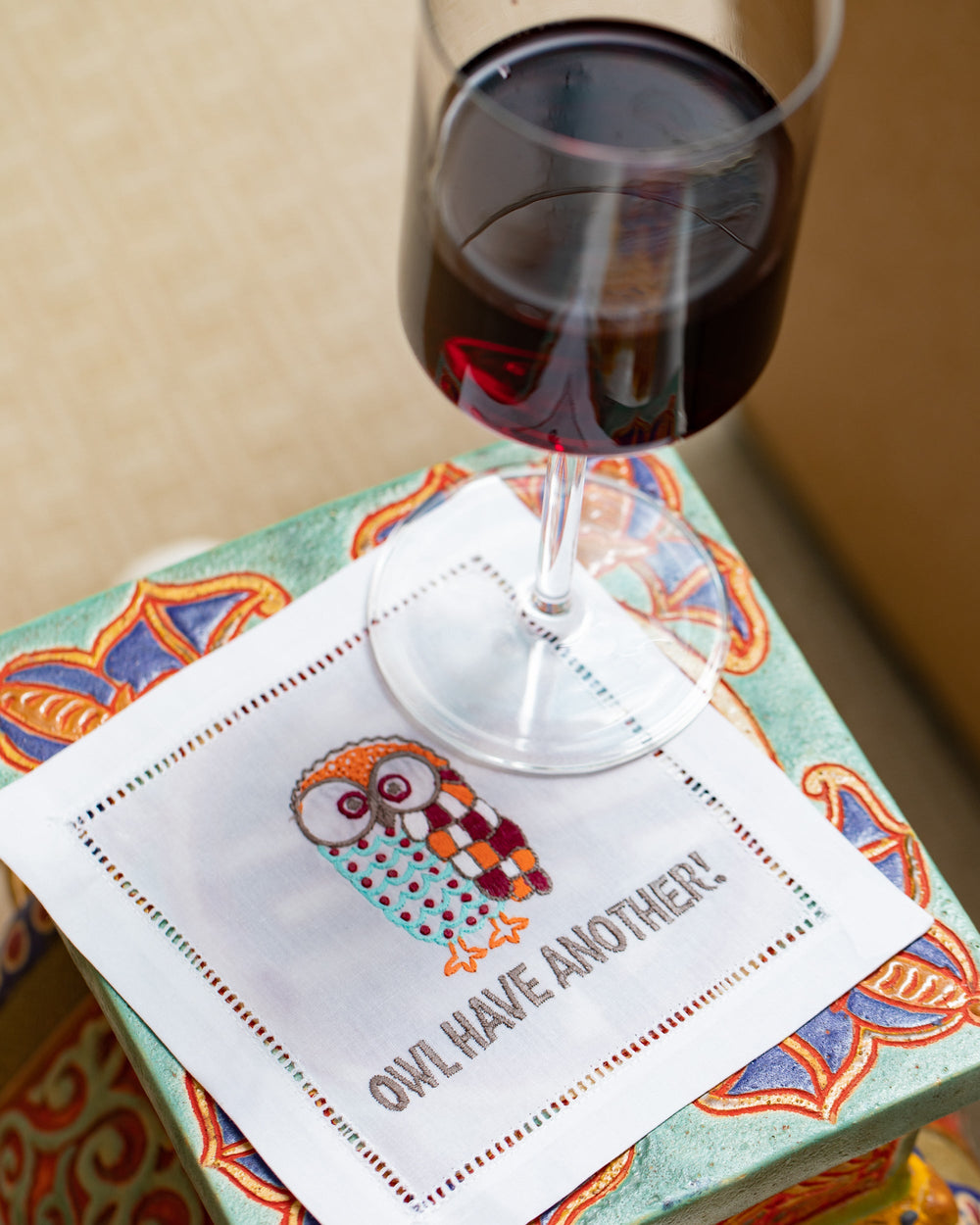 Top-down view of embroidered cocktail napkin with colorful owl and Owl Have Another! text, hemstitch edge on patterned box with wine glass