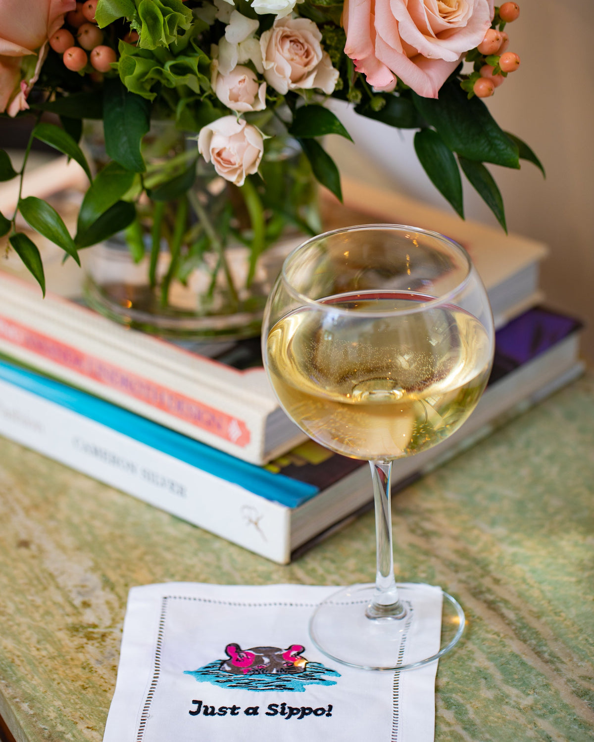 Close-up of Just a Sippo cocktail napkin with embroidered hippo and hemstitching beside a wine glass and stacked books