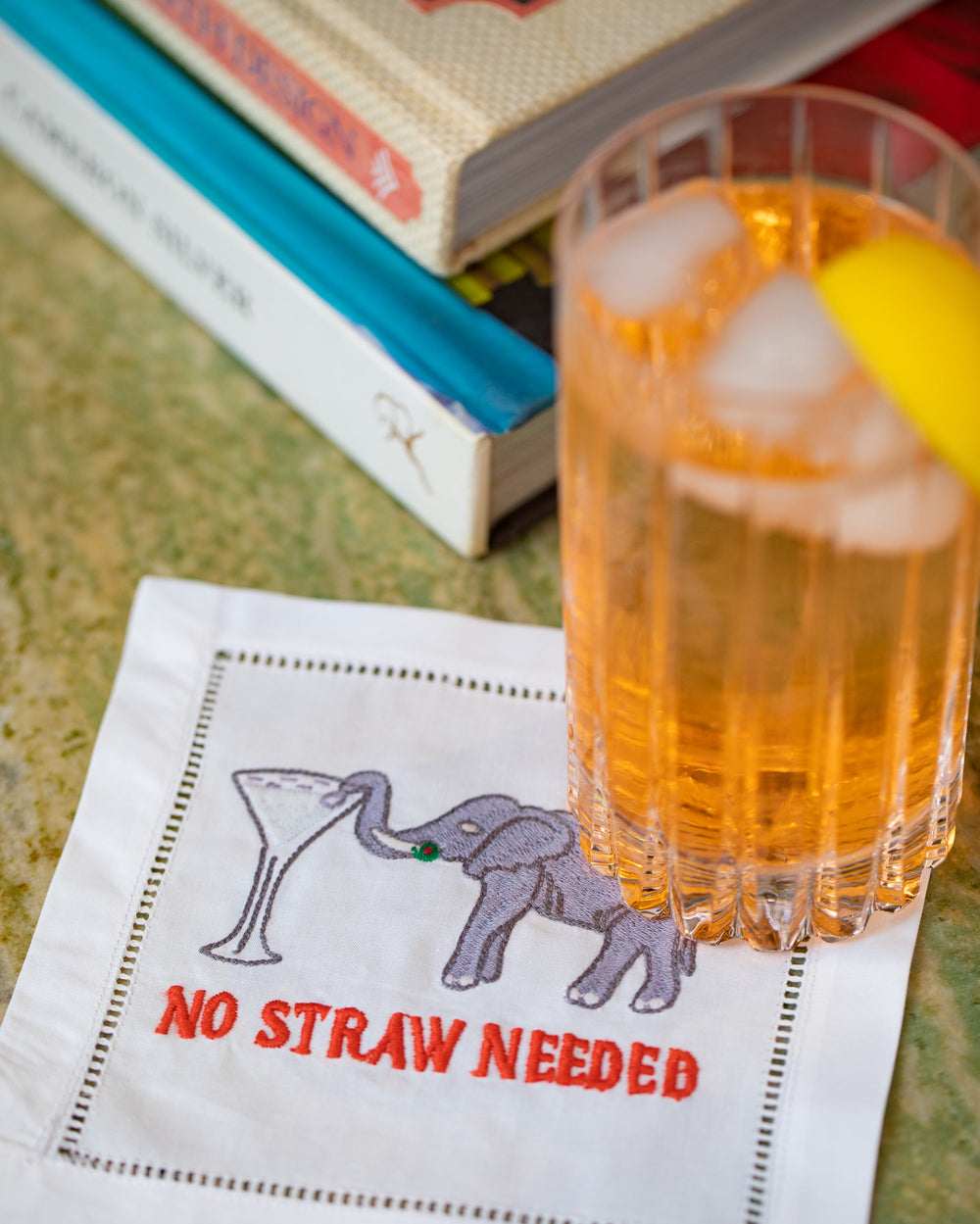 Close-up of No Straw Needed cocktail napkin showcasing embroidered elephant with martini glass and red “NO STRAW NEEDED” text beside an iced cocktail glass and stacked books