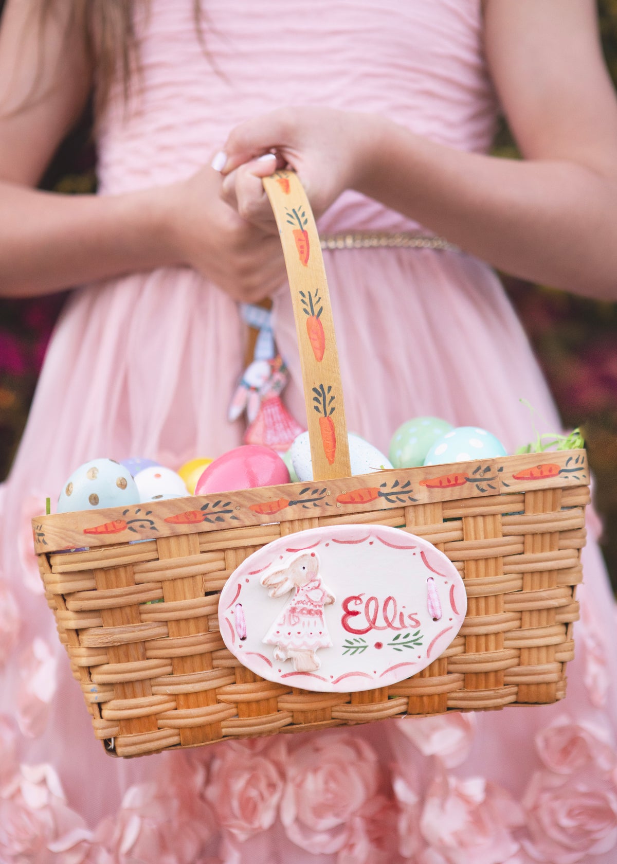 Close-up of hand-painted Easter basket with ceramic name plate Ellis, carrot-painted handle and pastel eggs held by a girl