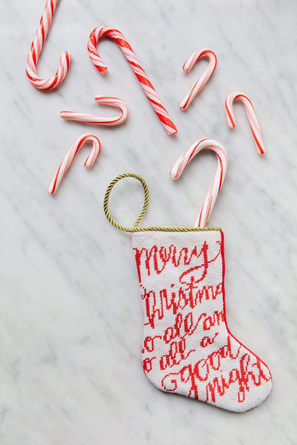 Top-down view of Merry Christmas bauble stocking showcasing red needlepoint script reading “Merry Christmas” and a gold hanging loop on a white marble background