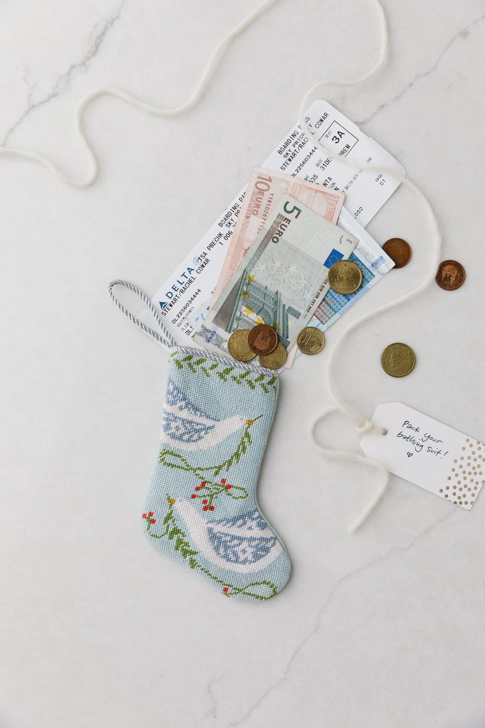 Top-down of Peace on Earth blue bauble stocking with needlepoint dove, euros, boarding pass, and gift tag on marble