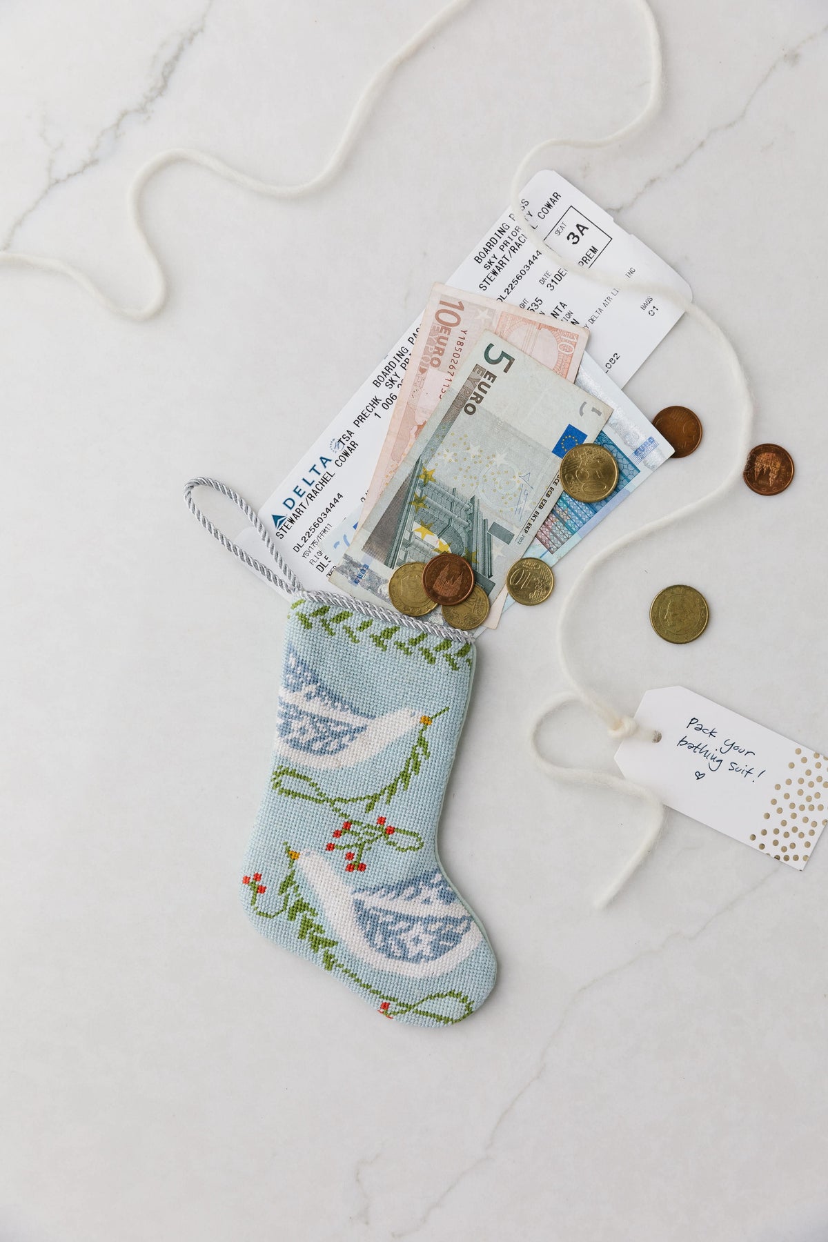 Top-down of Peace on Earth blue bauble stocking with needlepoint dove, euros, boarding pass, and gift tag on marble