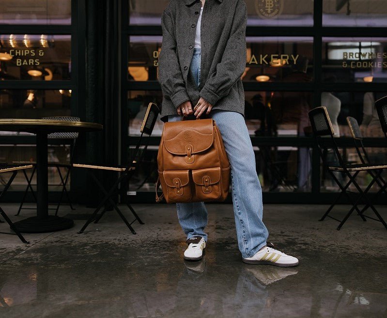 Person holding tan Blazer No. 278 leather backpack in front of a café, showing front pockets and top handle