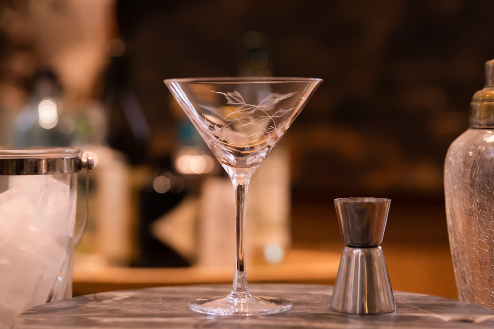 Front view of crystal martini glass showcasing hand-engraved fern design with blurred bar background