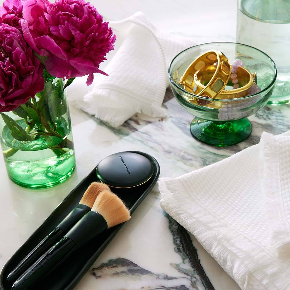 Green glass footed bowl holding bracelets on a marble tabletop with pink peonies and makeup brushes