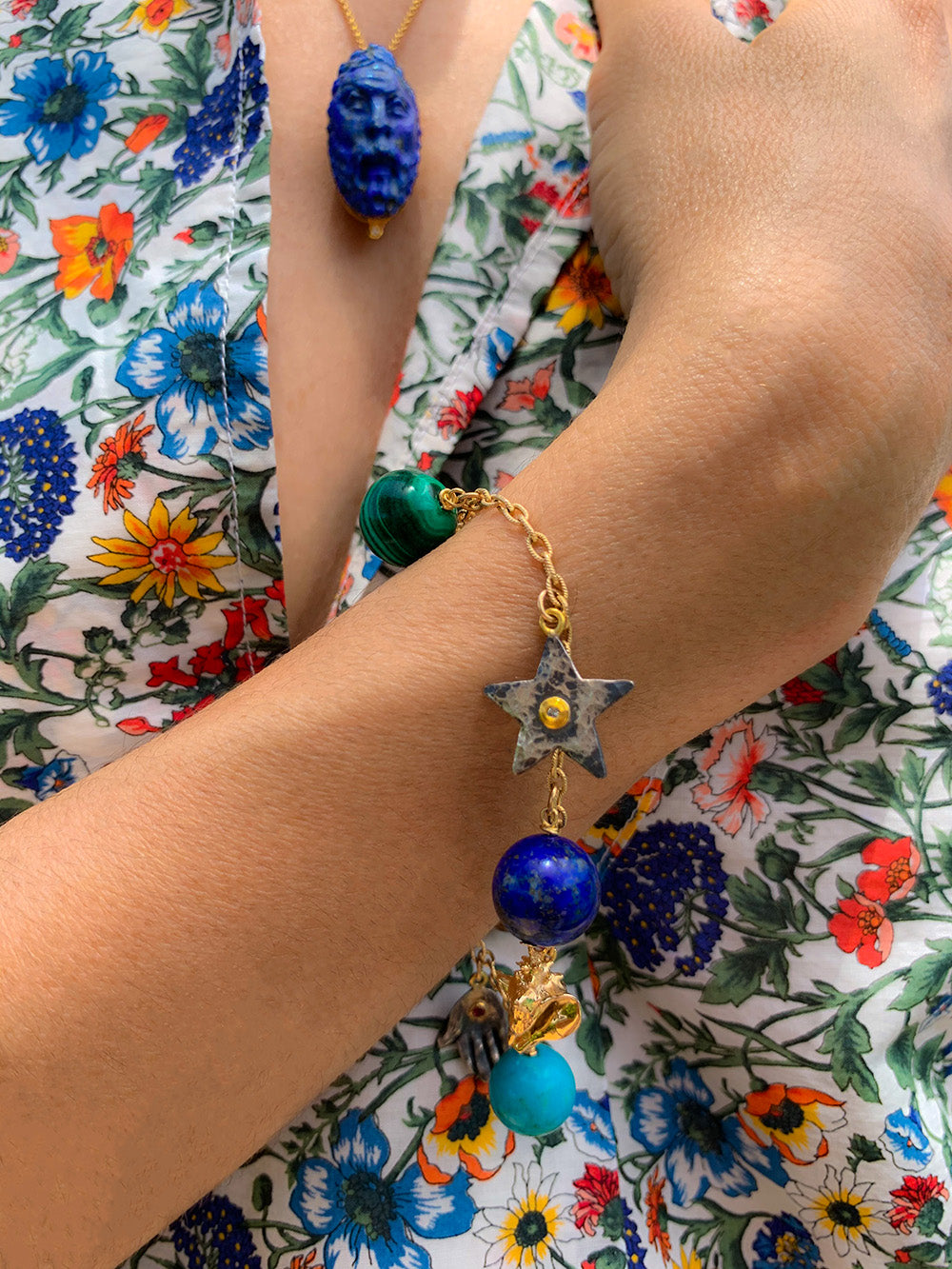 Close-up of Les Charmantes charm bracelet on wrist showing gold chain, star and blue-green gemstone charms on floral dress