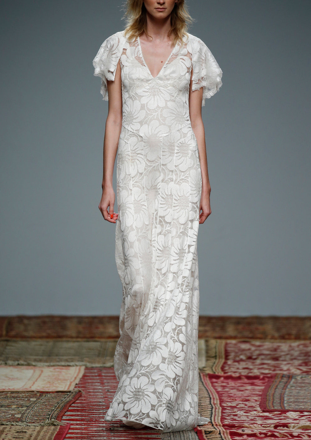 Front view of Dahlia Floral Lace Gown showcasing large floral lace pattern, V-neckline, flutter sleeves, and sheer full-length skirt on runway