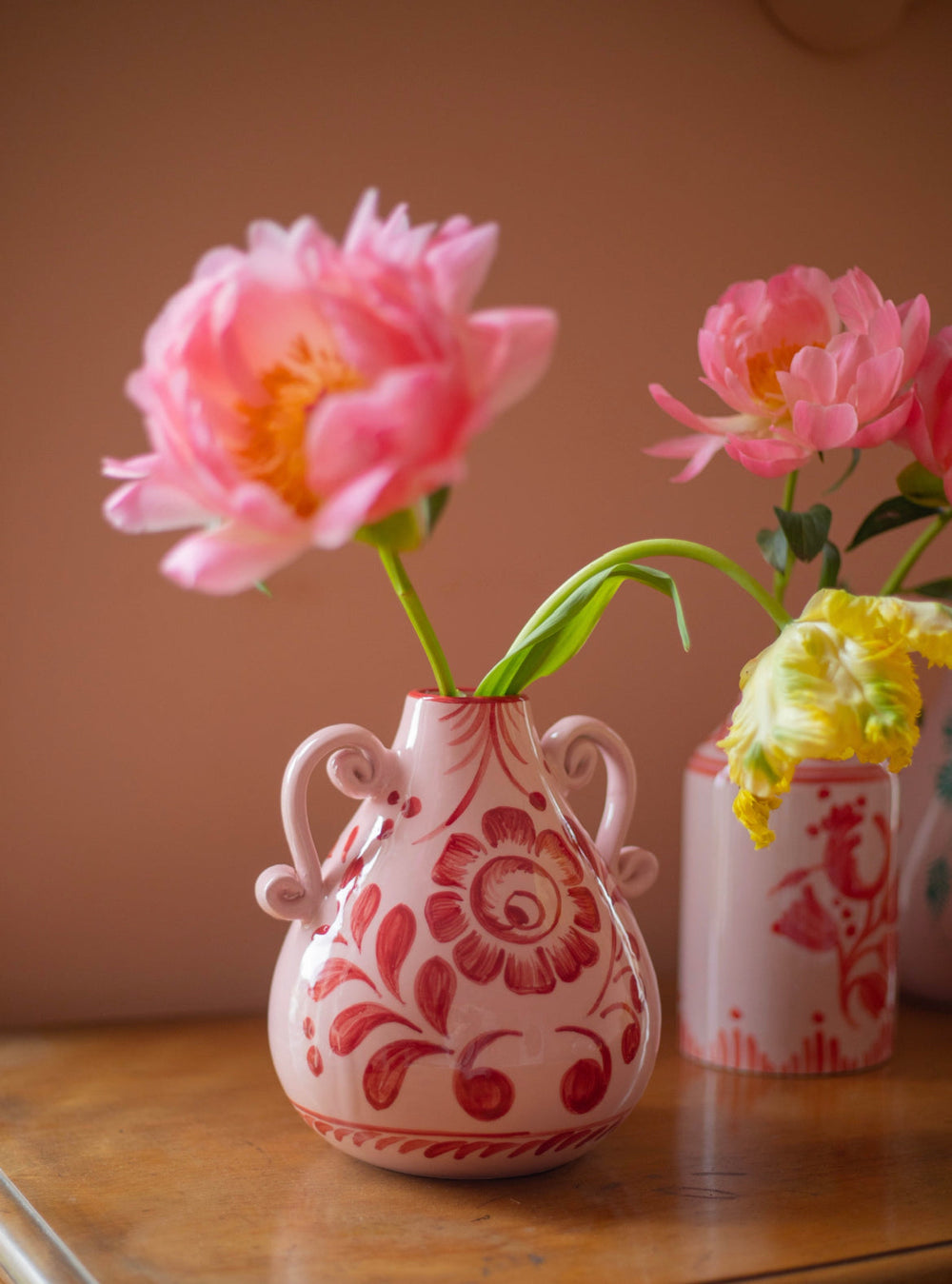 Front view of Amphora vase with hand-painted wine floral pattern and curled handles on wooden table against pink wall