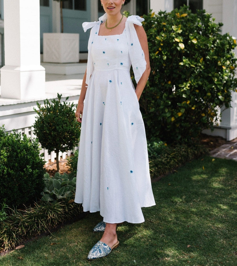 Three-quarter view of Sara Midi Dress in white linen with blue embroidered flowers and self-tie shoulder straps, worn outdoors