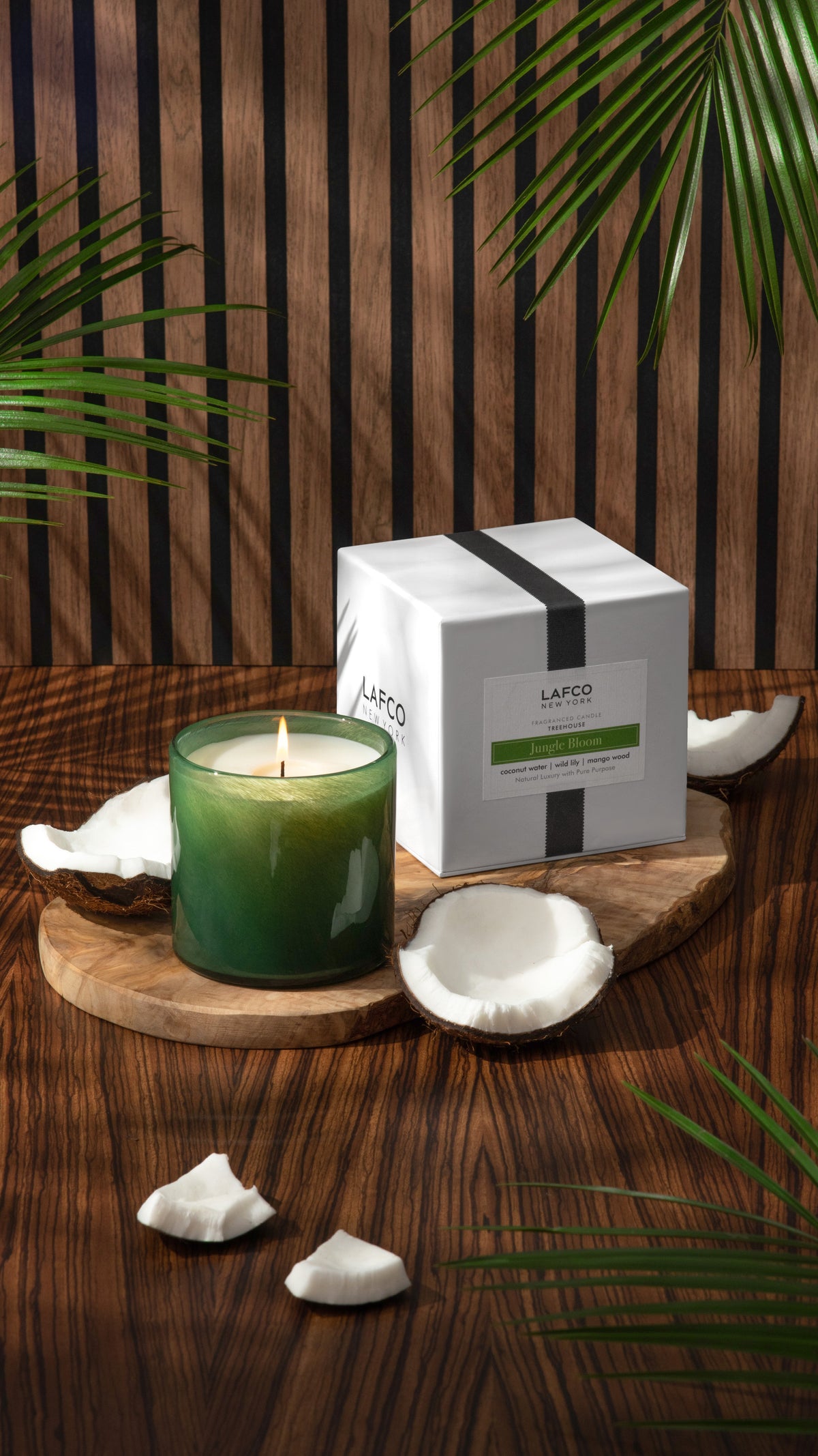 Angled view of Jungle Bloom candle in green glass, lit next to halved coconuts and white box on wood backdrop