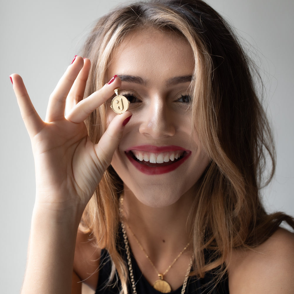 Young woman holding a gold coin pendant engraved with the letter J up to her eye, smiling with layered necklaces visible