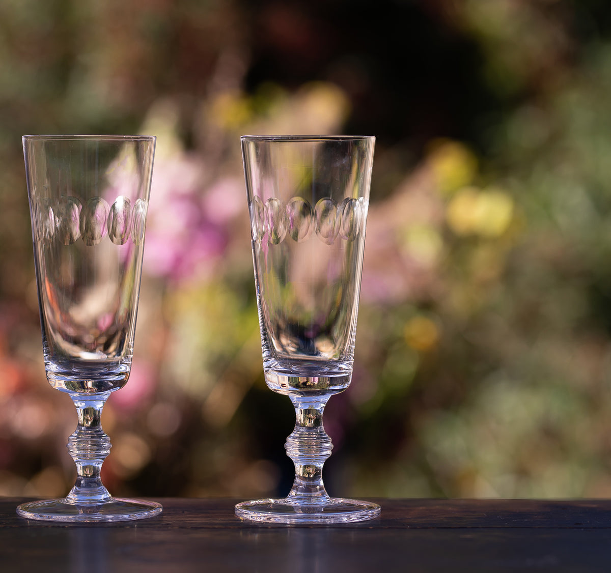 Front view of crystal champagne flutes with Lens Design showcasing engraved oval pattern and twisted stems against a floral backdrop