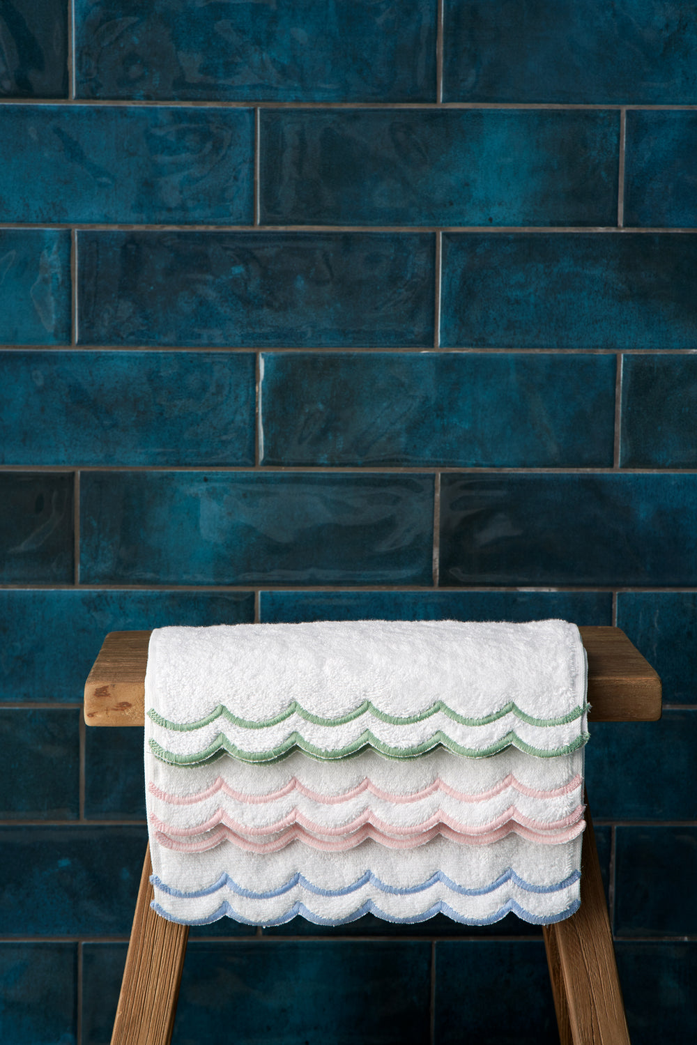 Folded Loulou Bath Mat on wooden stool showing scalloped embroidered edges in blue, pink, and green against dark teal tile