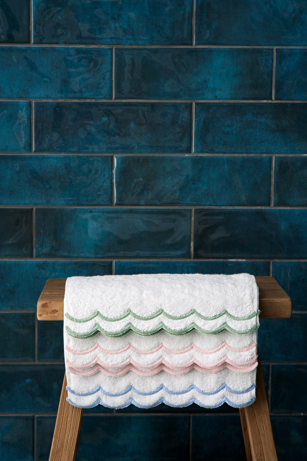 Folded Loulou Bath Mat on wooden stool showing scalloped embroidered edges in blue, pink, and green against dark teal tile