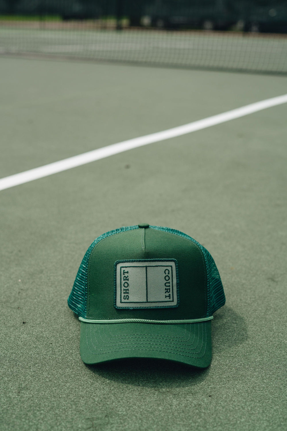Front view of Short Court trucker hat, green with logo patch reading SHORT COURT, braided trim and mesh back on tennis court