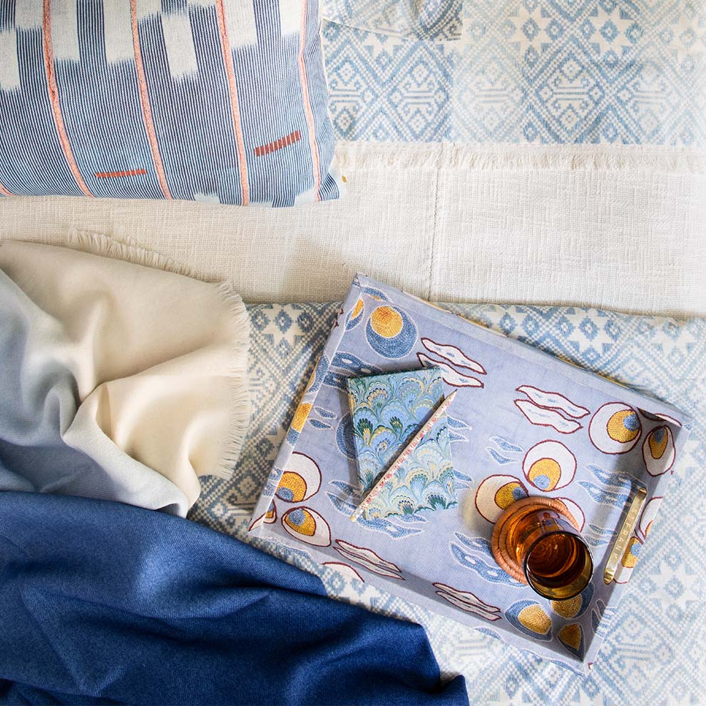 Overhead view of blue chambray pillow on layered throws with a patterned tray holding a folded napkin and amber glass