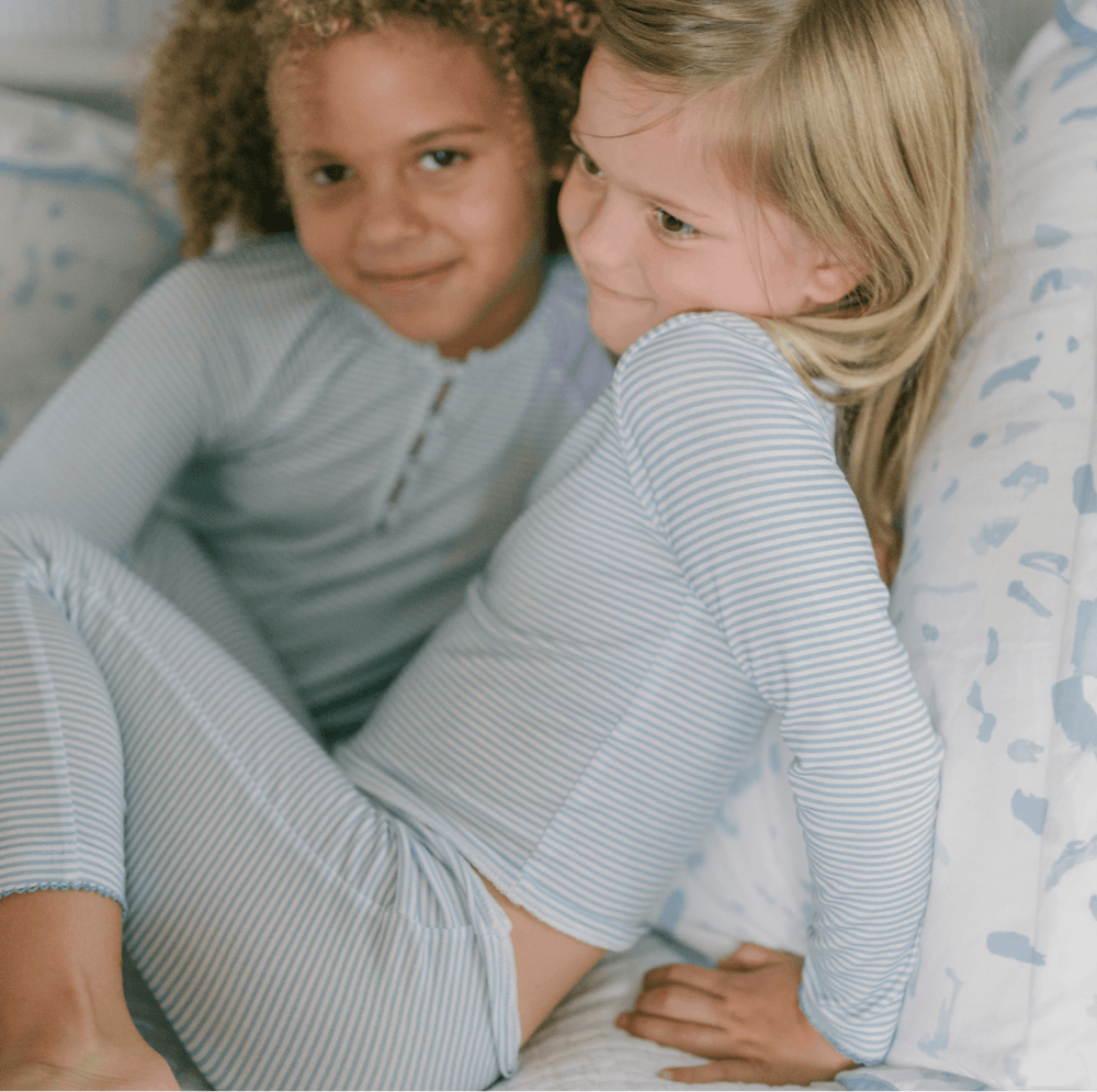 Two girls wearing powder blue striped Pima pajama sets sitting and smiling on a bed with patterned pillows