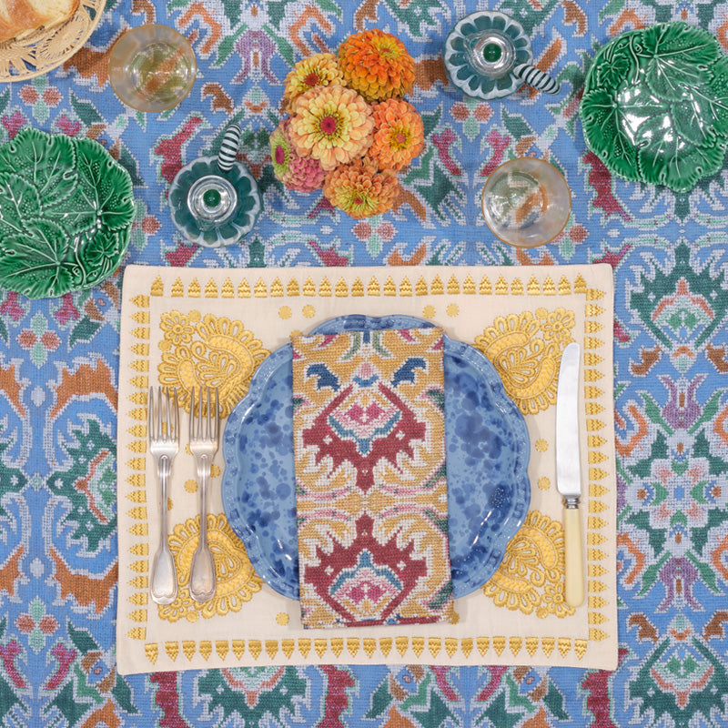 Blue speckled scalloped dinner plate with patterned napkin on cream placemat over blue ikat cloth, with silverware and flowers