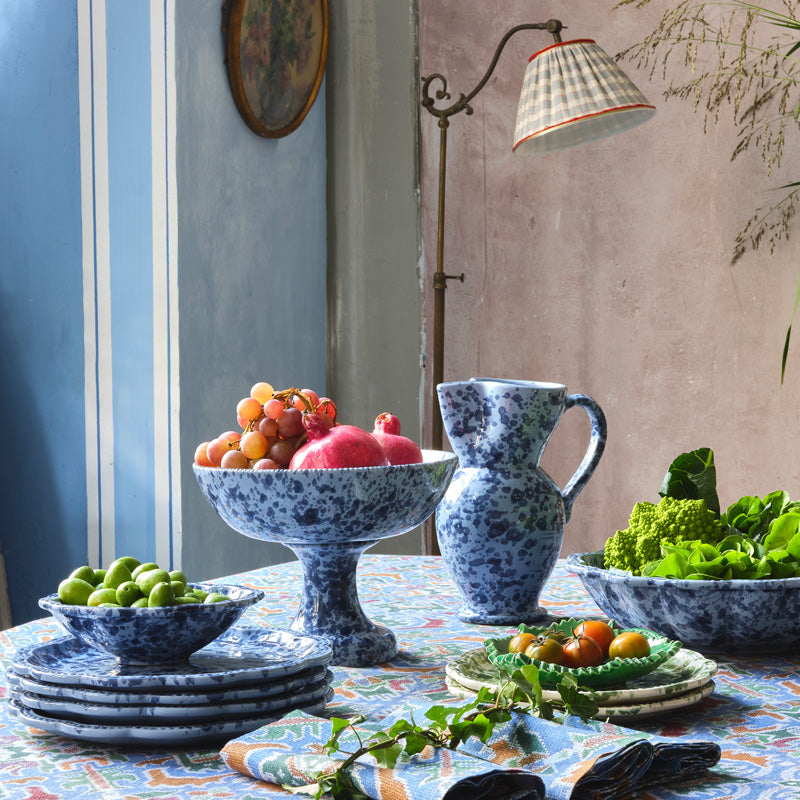 Front view of Speckled Fruit Stand in Blue showing hand-painted finish, scalloped edge, and fruit on a patterned table