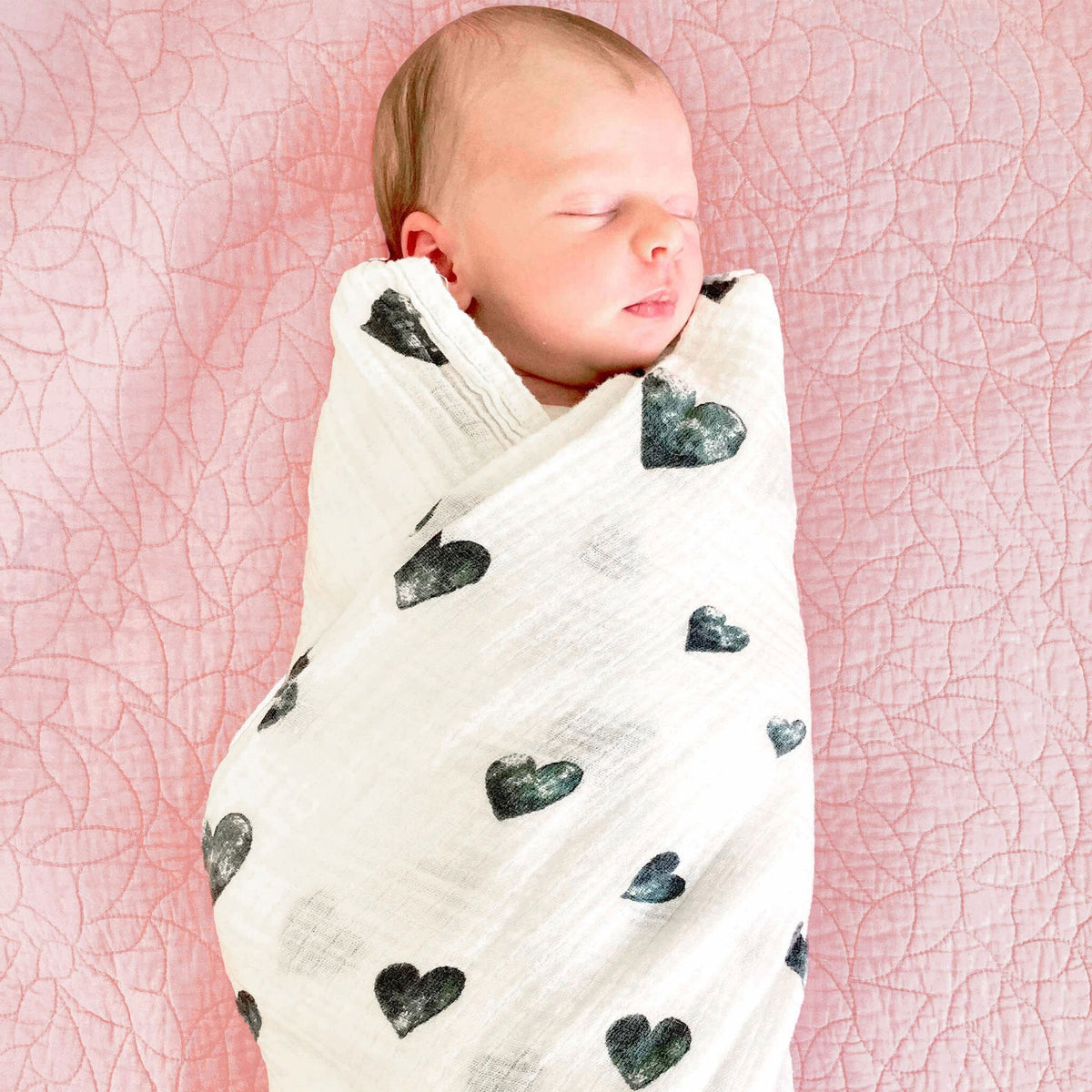 Top-down view of organic swaddle blanket swaddling a newborn, white muslin with black heart print on a pink quilt