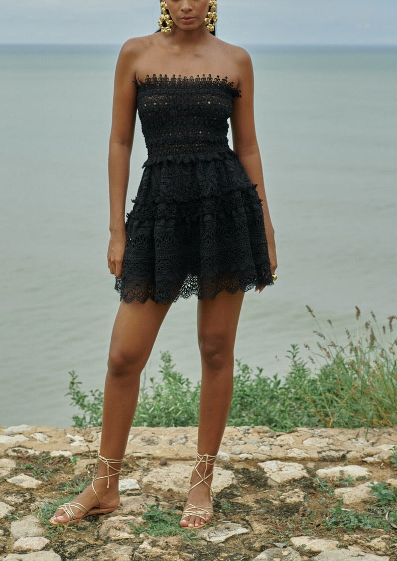 Front view of Waimari Vallarta strapless black mini dress showcasing tiered Guipure lace and scalloped hem on a seaside path