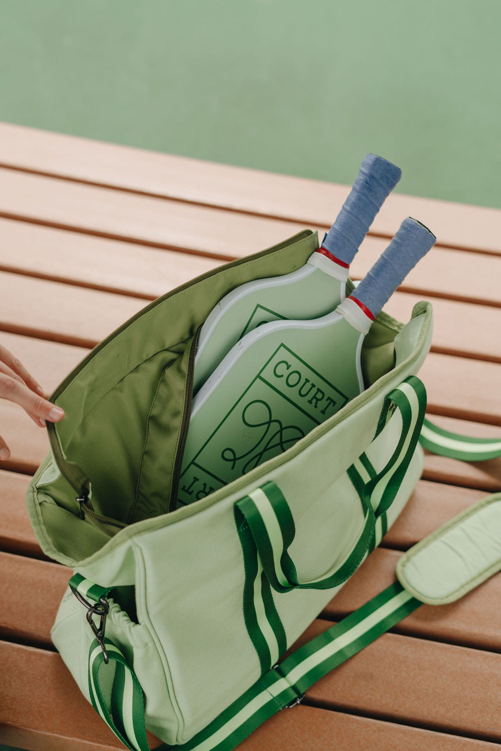 Top-down view of a light green Courtside doubles tote showing two green pickleball paddles with blue grips inside and a green striped shoulder strap on a wooden bench