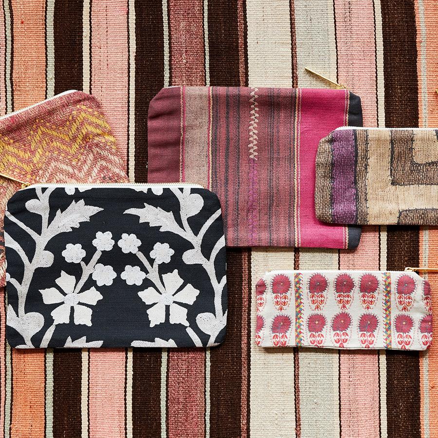 Front view of Fuchsia Daisy Suzani slim zip pouch showcasing its pink floral pattern and gold zipper among assorted pouches on striped textile